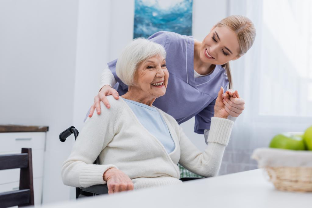 glimlachende verpleegster hand in hand met vrolijke gehandicapte vrouw thuis - Foto, afbeelding