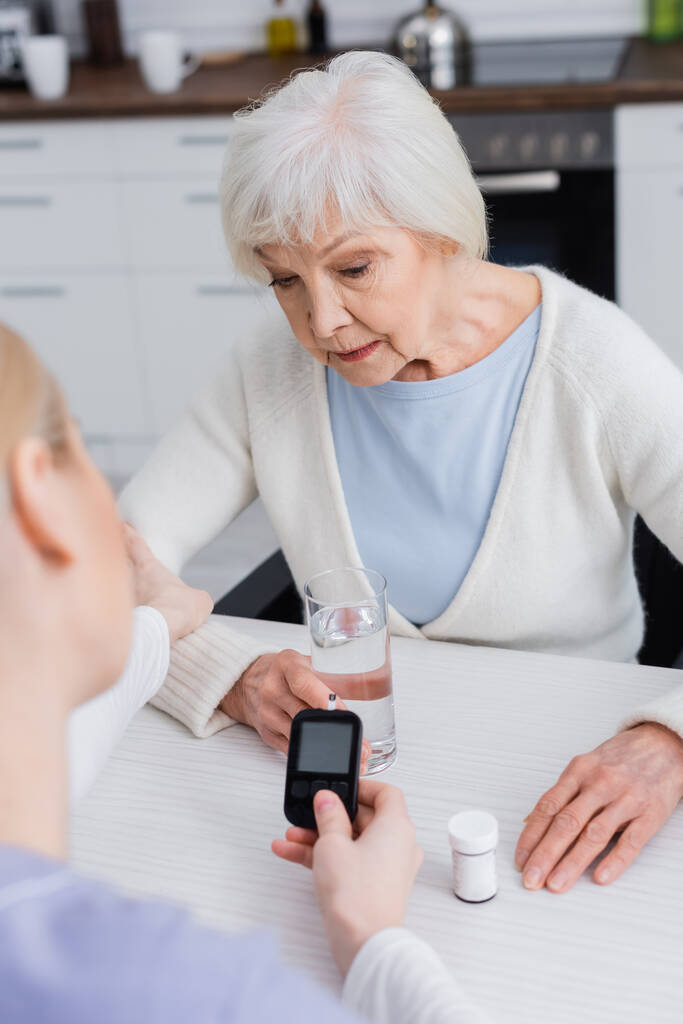 Hemşire yaşlı diyabetik kadının yanında glukometre tutuyor ve ilaç kutusu bulanık ön planda. - Fotoğraf, Görsel