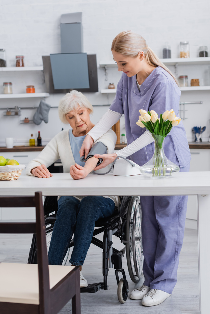 enfermera joven que mide la presión arterial de una mujer discapacitada en silla de ruedas en casa - Foto, Imagen
