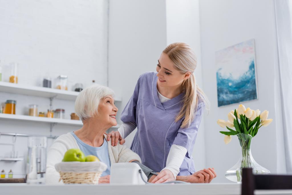 infermiera sorridente guardando felice donna anziana mentre misura la pressione sanguigna in cucina - Foto, immagini