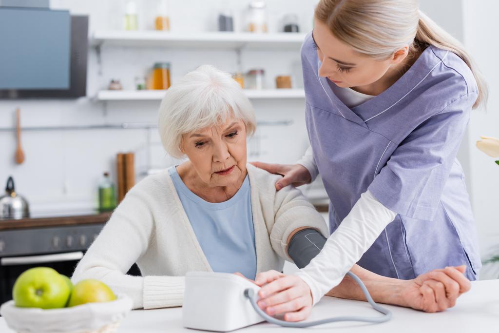 jovem enfermeira tocando no ombro da mulher idosa enquanto mede sua pressão arterial - Foto, Imagem