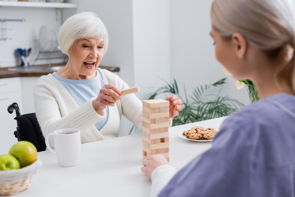 eccitata donna anziana giocare torre di legno gioco con assistente sociale in primo piano sfocato - Foto, immagini