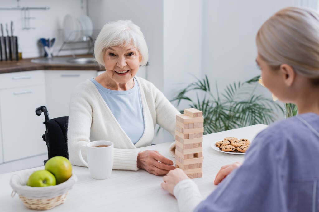 Yaşlı engelli kadın hemşireyle tahta bloklar oynarken kameraya gülümsüyor - Fotoğraf, Görsel