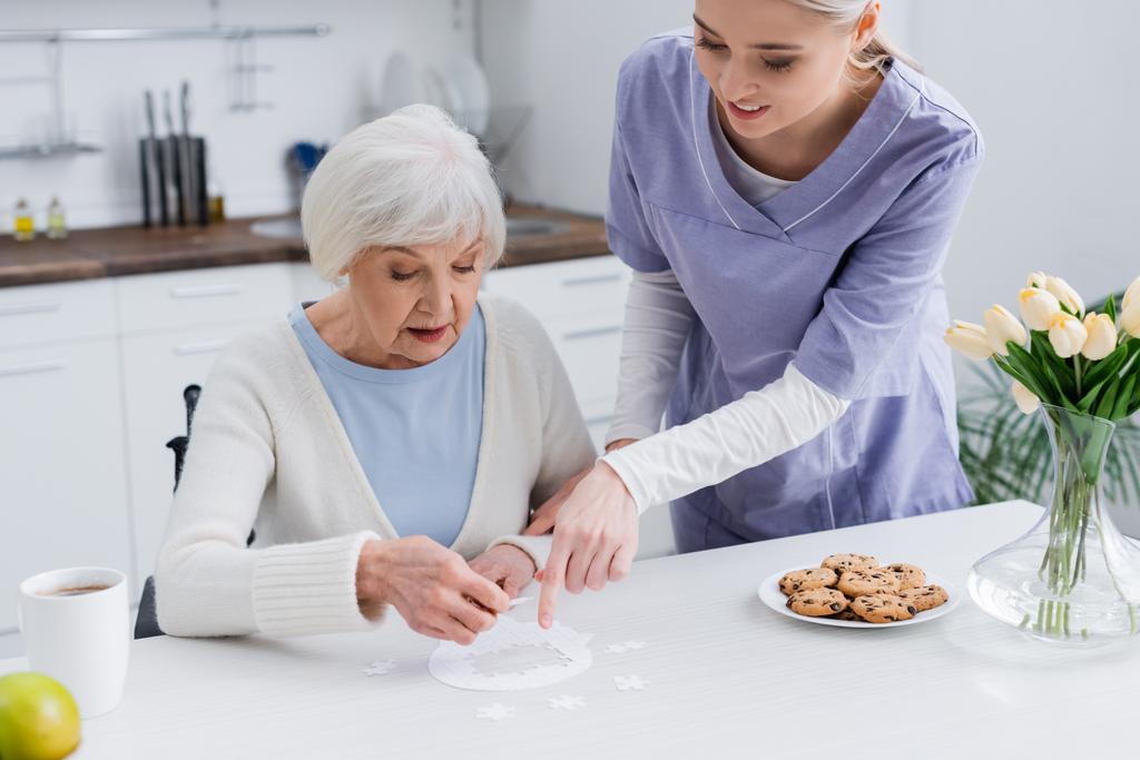 joven enfermera señalando rompecabezas cerca de anciana mujer en la cocina - Foto, Imagen