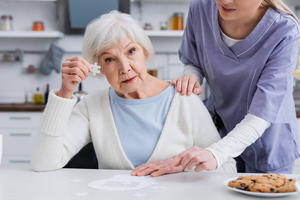ηλικιωμένη γυναίκα δείχνει κομμάτι του παζλ, ενώ νοσοκόμα αγγίζοντας τον ώμο της - Φωτογραφία, εικόνα