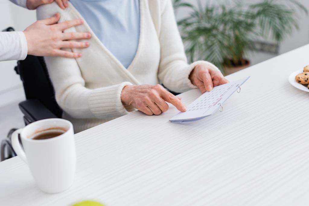 Ausgeschnittene Ansicht der Krankenschwester berührt Schulter der Seniorin, die auf Kalender zeigt, während sie an Gedächtnisverlust leidet - Foto, Bild