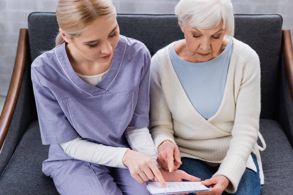 young nurse and senior woman, suffering from memory loss, pointing at calendar while sitting on couch - Photo, Image