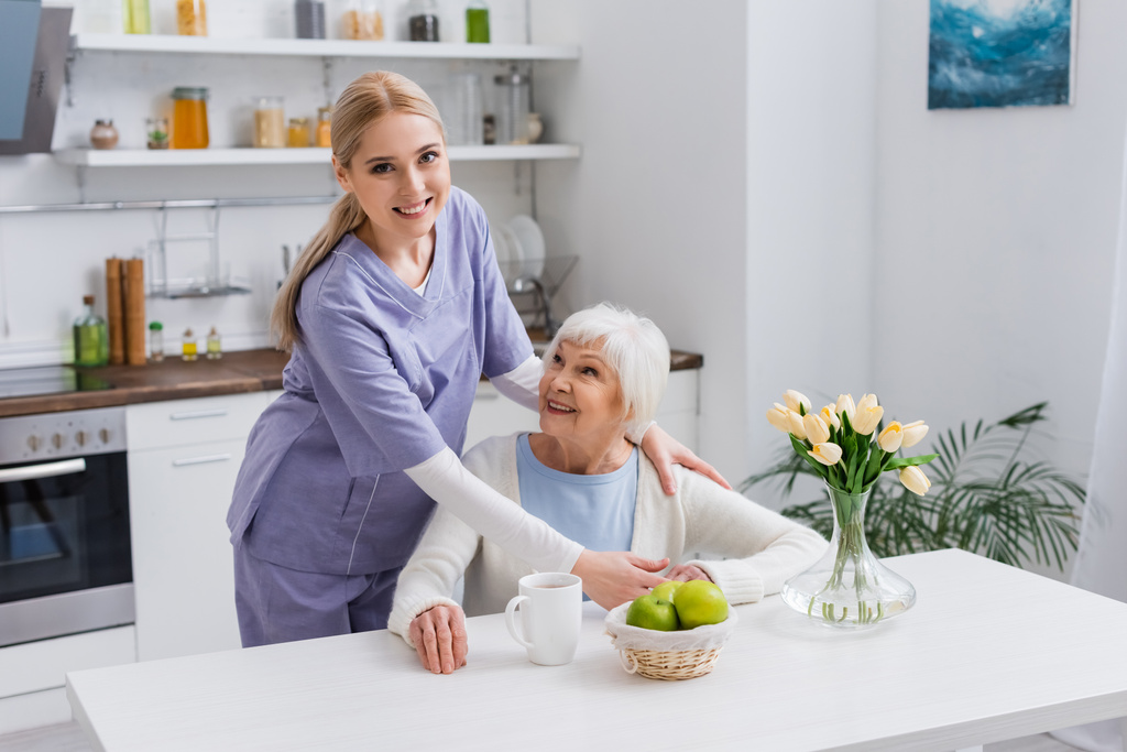 joven enfermera sonriendo a cámara mientras abrazando feliz anciano mujer en cocina - Foto, imagen