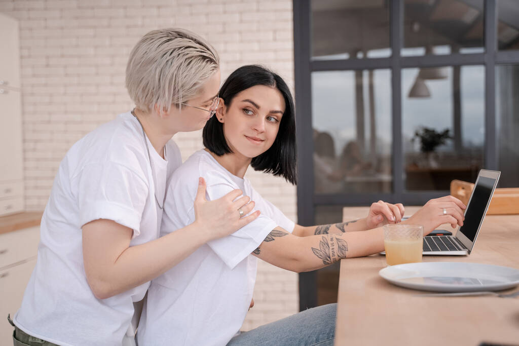 junge Frau schaut tätowierte Freundin mit Laptop in Küche an  - Foto, Bild