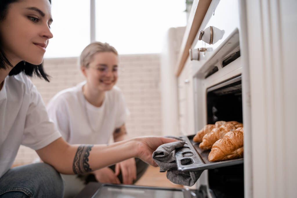 tätowiertes lesbisches Paar blickt in Küche auf Backblech mit frisch gebackenen Croissants  - Foto, Bild