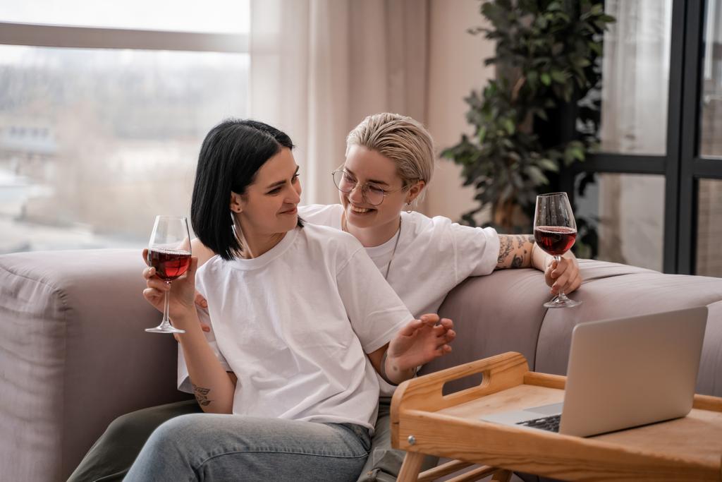feliz pareja lesbiana viendo la película en el ordenador portátil mientras sostiene vasos de vino tinto  - Foto, imagen