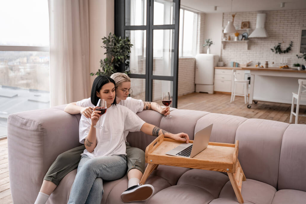 молодая лесбийская пара смотрит кино на ноутбуке, держа бокалы красного вина в гостиной - Фото, изображение