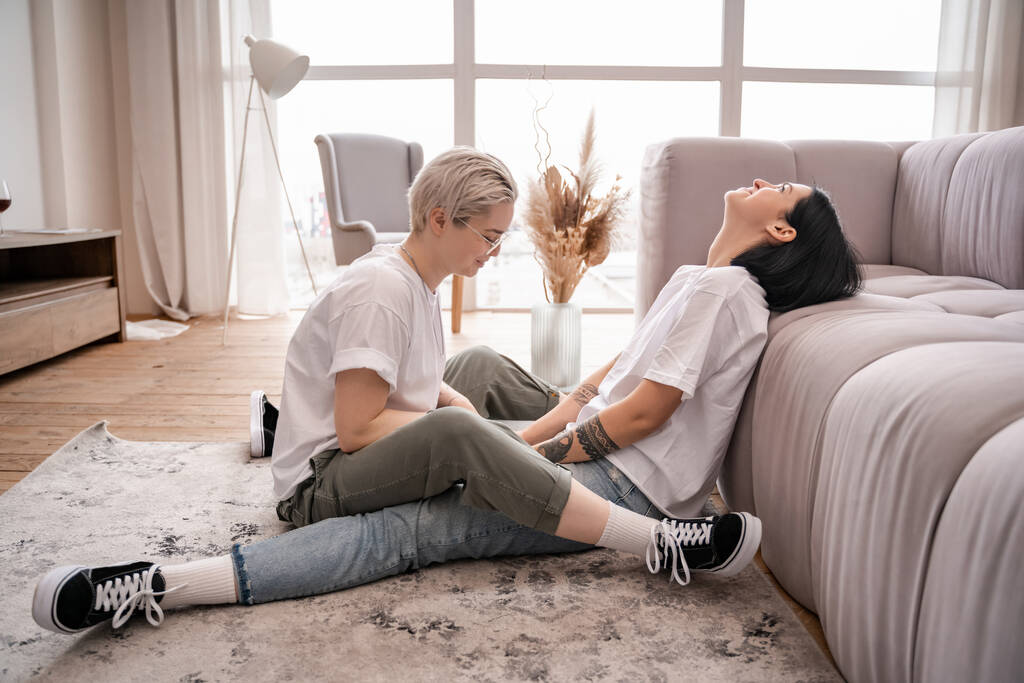 Tätowierte Frau sitzt mit brünetter Freundin auf Teppich in der Nähe von Couch  - Foto, Bild