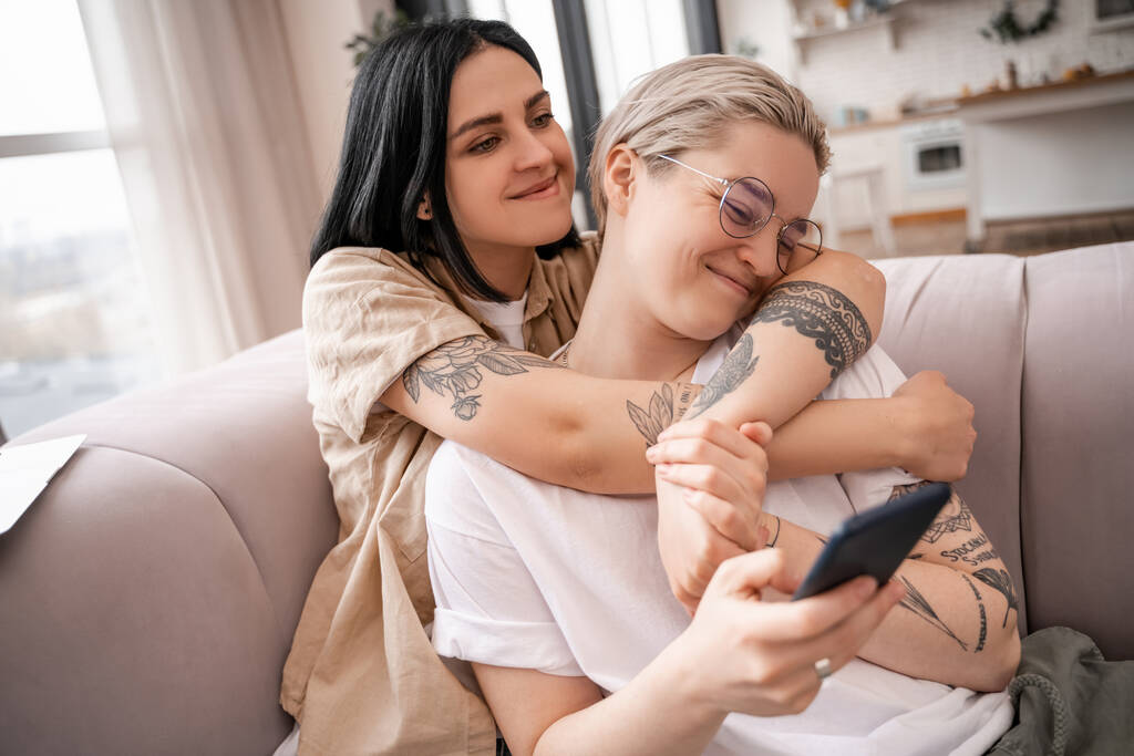 lesbian couple hugging while sitting on couch and using smartphone  - Photo, Image