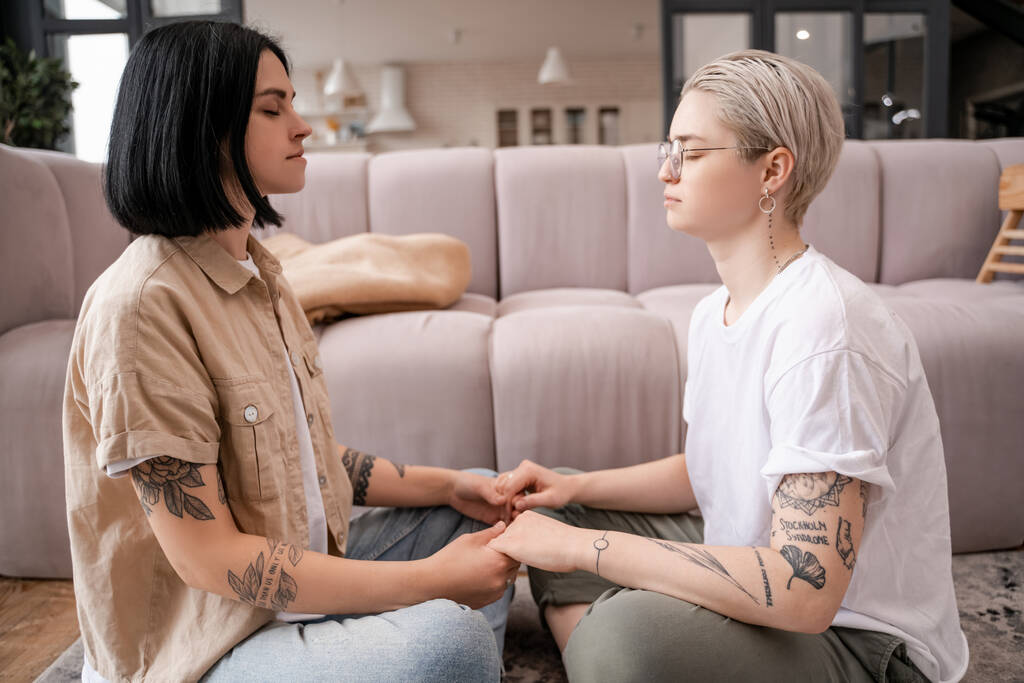 vista laterale della coppia lesbica seduta e tenuta per mano mentre medita in soggiorno  - Foto, immagini