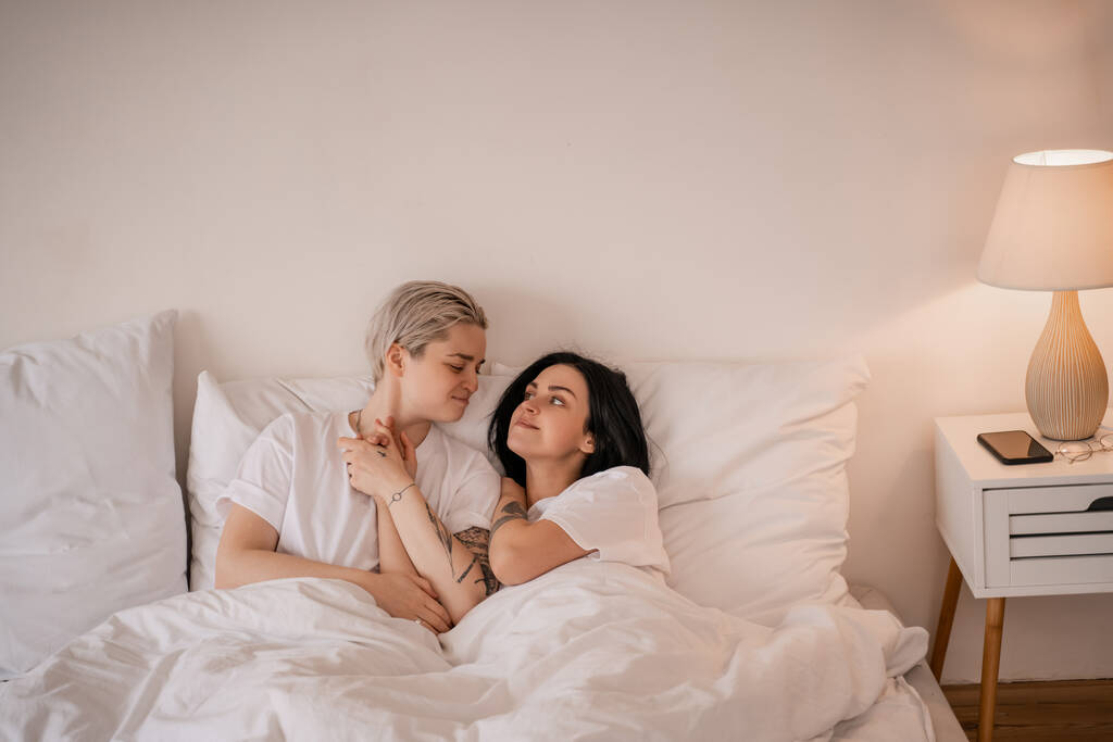 usmívající se lesbický pár ležící v posteli a držící se za ruce - Fotografie, Obrázek