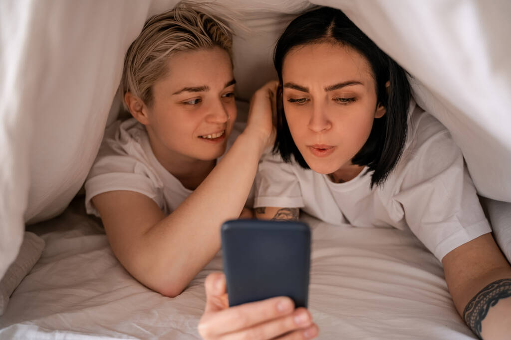 彼女と毛布の下に横たわっている間にスマートフォンでテキストメッセージを送る若い女性  - 写真・画像