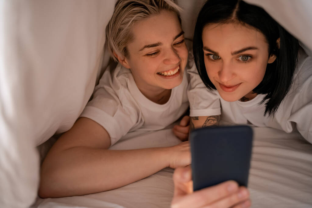 молодая женщина сообщения на смартфоне во время лежания под одеялом с подругой  - Фото, изображение