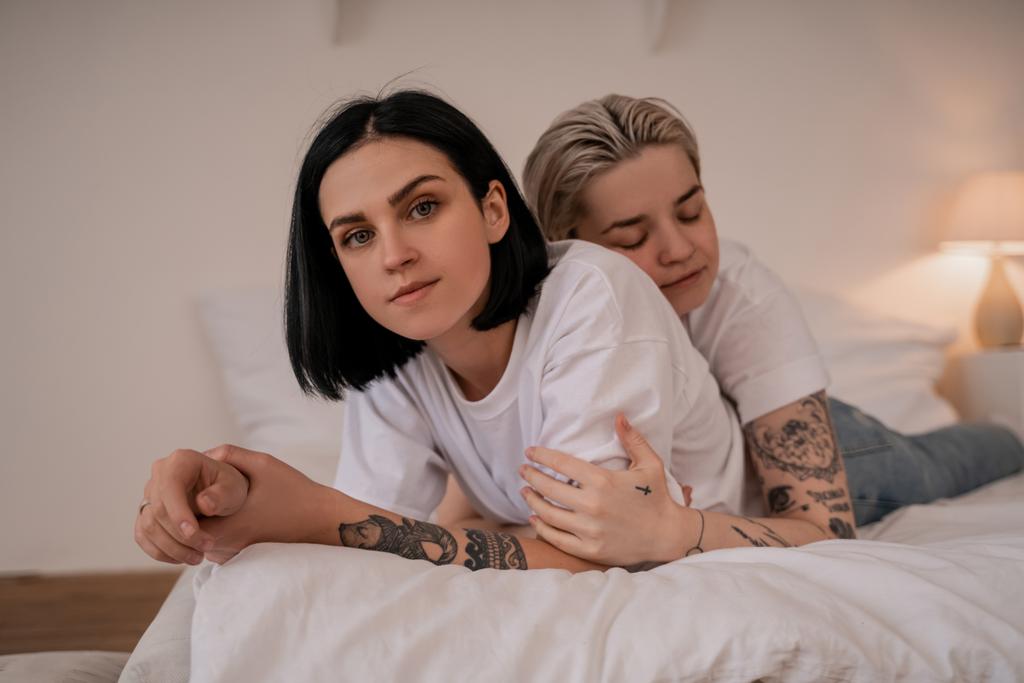 Genç lezbiyen kadın dövmeli kız arkadaşına sarılıyor ve yatakta uzanıyor. - Fotoğraf, Görsel