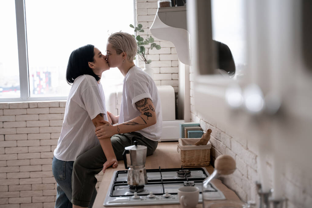 zijaanzicht van jonge vrouw zitten op keukentafel en zoenen met vriendin in de buurt koffiepot op fornuis  - Foto, afbeelding