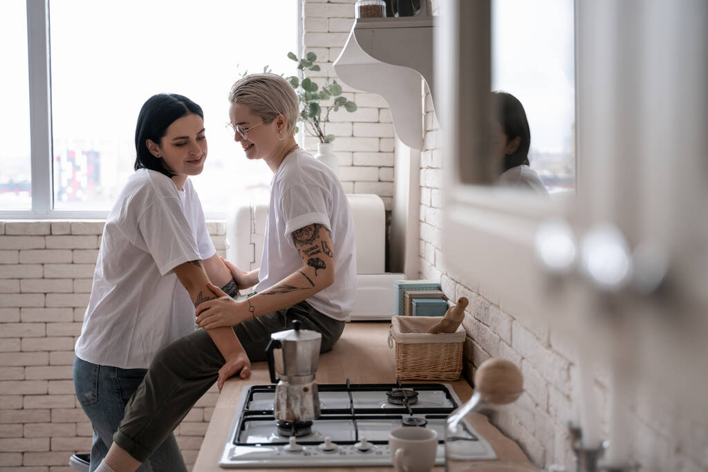 happy young woman sitting on kitchen table and hugging with girlfriend near coffee pot on stove  - Photo, Image