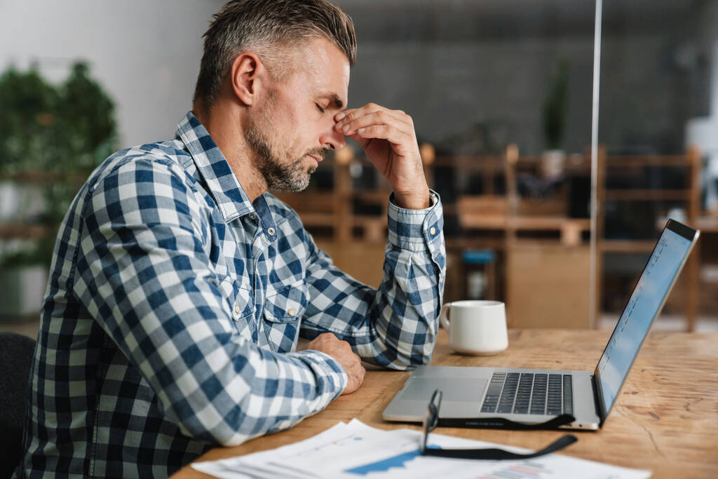 Hombre gris infeliz con dolor de cabeza trabajando con el ordenador portátil mientras está sentado en la oficina - Foto, Imagen