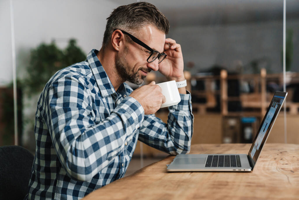 Blij ongeschoren man drinken van koffie tijdens het werken met laptop op kantoor - Foto, afbeelding