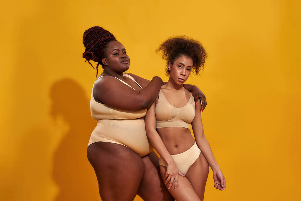 Dos mujeres afroamericanas serias en ropa interior beige con diferentes tamaños de cuerpo de pie juntas, posando aisladas sobre fondo amarillo - Foto, imagen