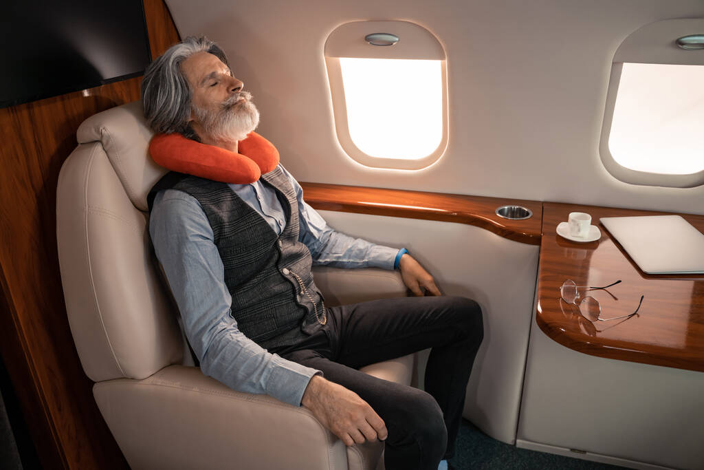 Зрелый бизнесмен в подушке для шеи спит рядом с ноутбуком и кофе в частном самолете  - Фото, изображение