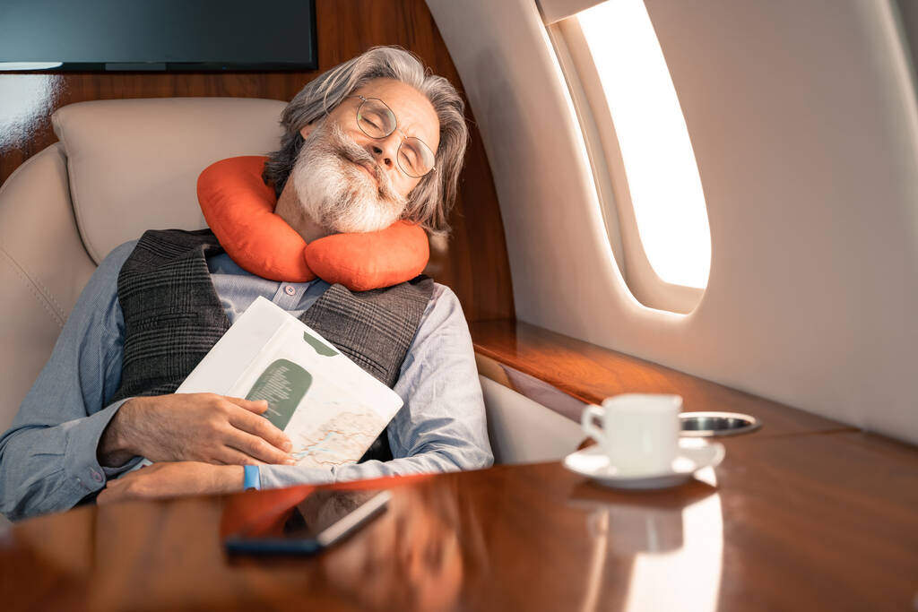 Επιχειρηματίας στο μαξιλάρι λαιμό περιοδικό κρατώντας ενώ κοιμάται κοντά σε καφέ και smartphone στο αεροπλάνο  - Φωτογραφία, εικόνα