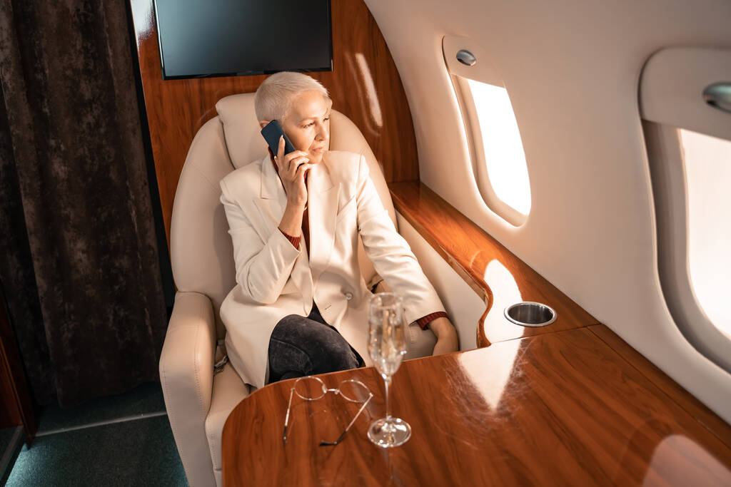 Зрелая деловая женщина разговаривает по смартфону возле размытого шампанского и очков в самолете  - Фото, изображение