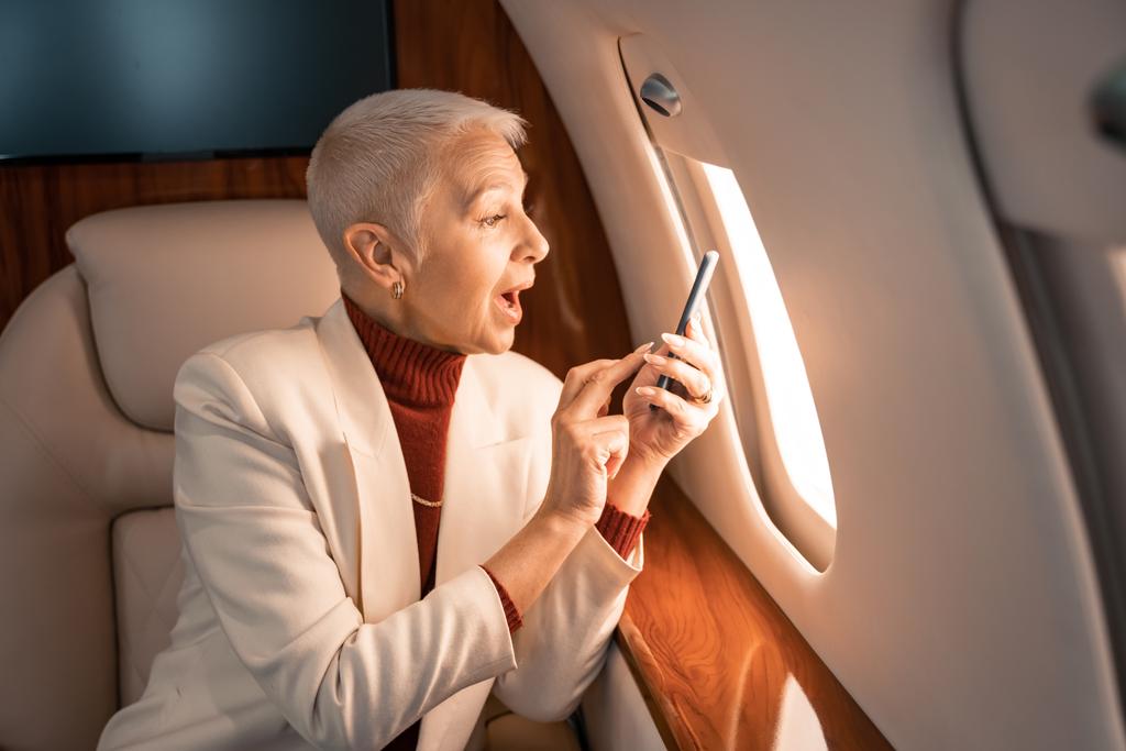 Erstaunte Geschäftsfrau mit Smartphone blickt im Privatflugzeug auf Fenster  - Foto, Bild