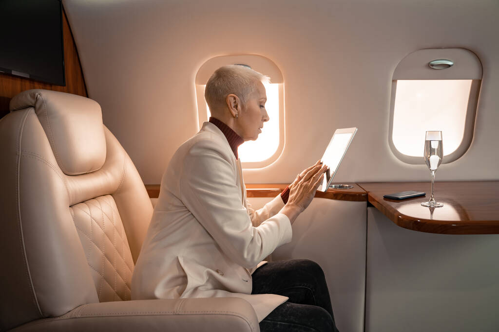 Вид збоку зрілої бізнес-леді, використовуючи цифровий планшет біля смартфона і келиха шампанського в літаку
 - Фото, зображення