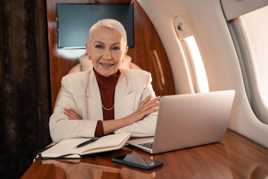 Usmívající se podnikatelka sedí v blízkosti zařízení a notebooku v letadle  - Fotografie, Obrázek