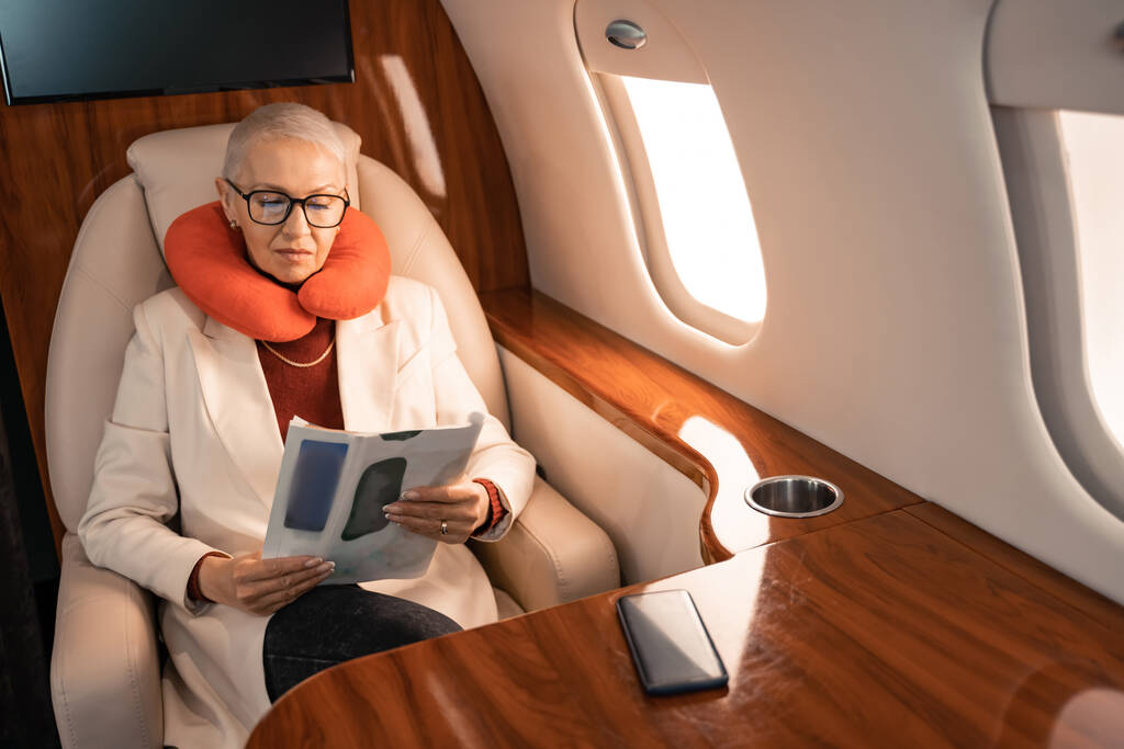 Zakenvrouw in de nek kussen lezen tijdschrift in de buurt wazig smartphone in het vliegtuig  - Foto, afbeelding