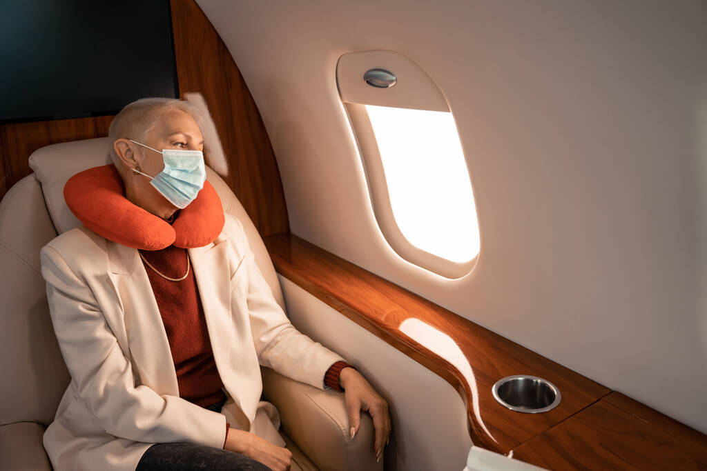 Üzletasszony nyakpárnában és orvosi maszkban nézi a repülőgép ablakát.  - Fotó, kép