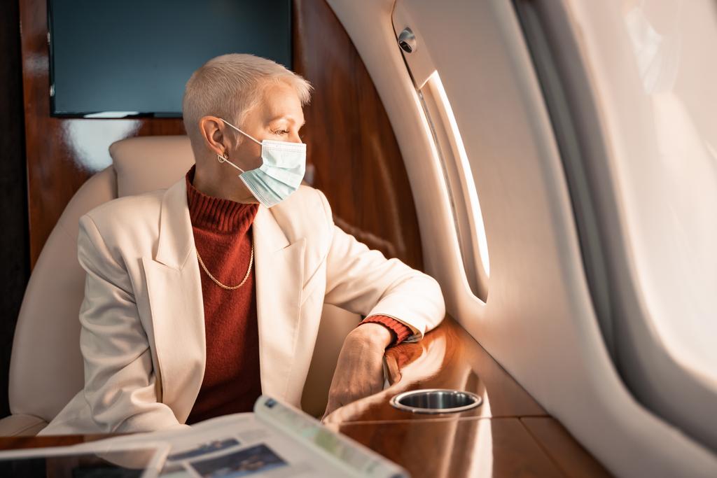 Geschäftsfrau mit medizinischer Maske blickt auf Flugzeugfenster neben Magazin und digitalem Tablet - Foto, Bild
