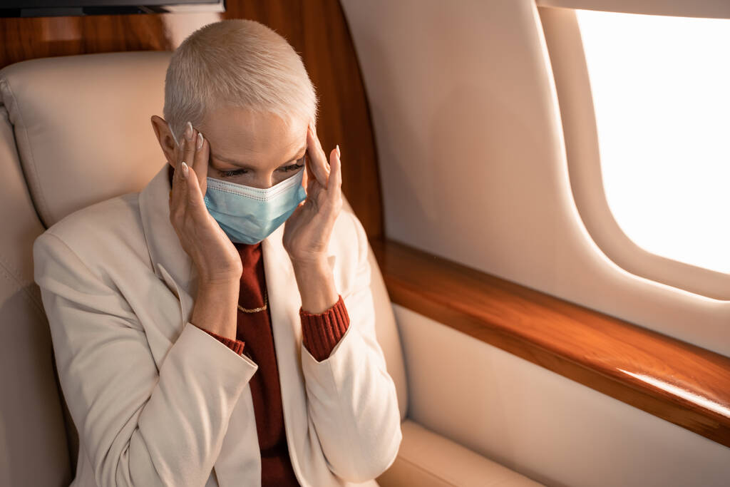 Деловая женщина в медицинской маске страдает от головной боли в самолете  - Фото, изображение