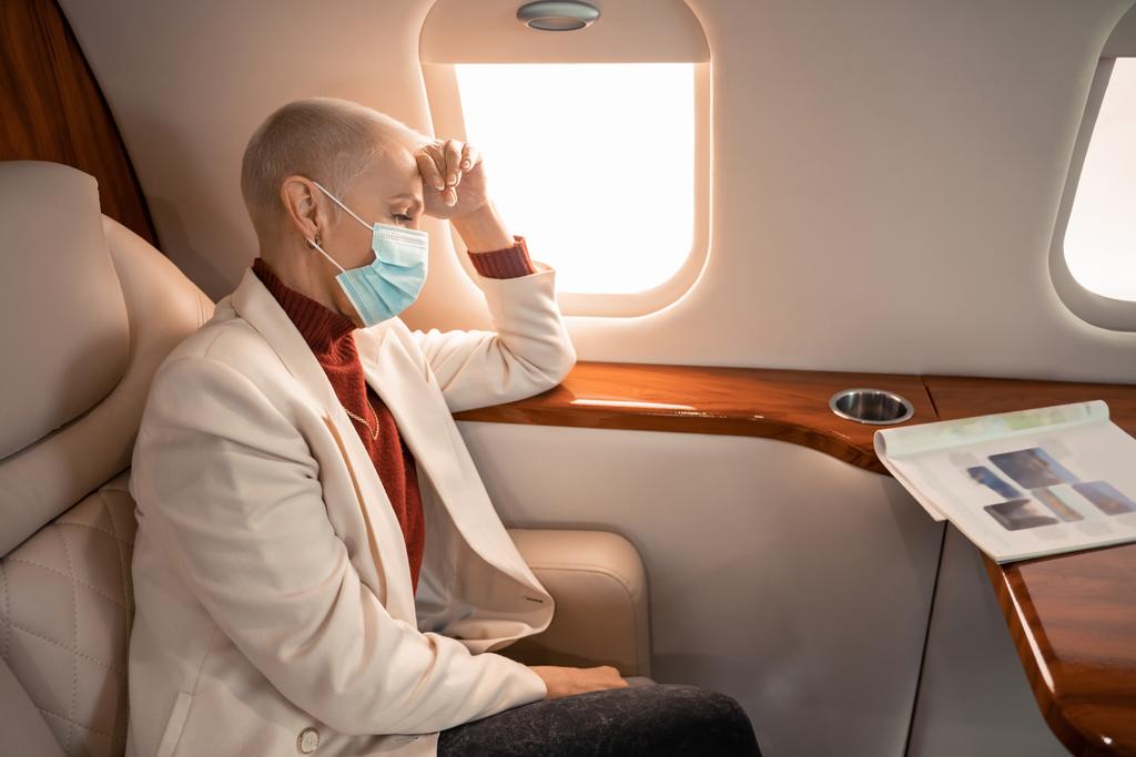 Επιχειρηματίας με ιατρική μάσκα που κοιτάζει περιοδικό σε ιδιωτικό αεροπλάνο  - Φωτογραφία, εικόνα