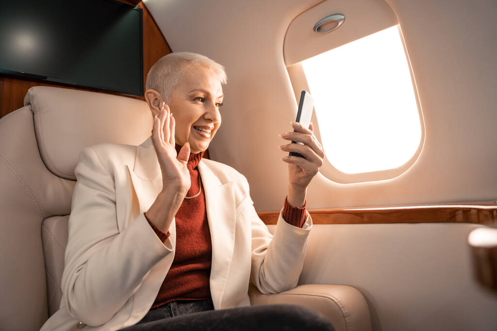 Glimlachende zakenvrouw zwaaiende hand tijdens videogesprek op smartphone in het vliegtuig  - Foto, afbeelding