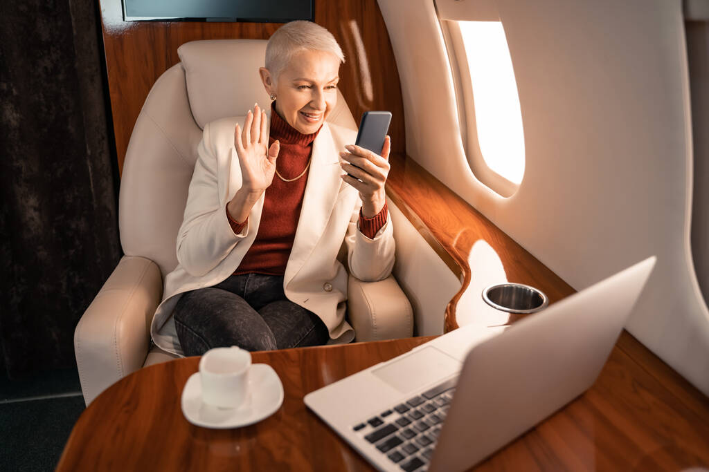 Mujer de negocios que tiene videollamada en el teléfono inteligente cerca de café y portátil en jet  - Foto, Imagen