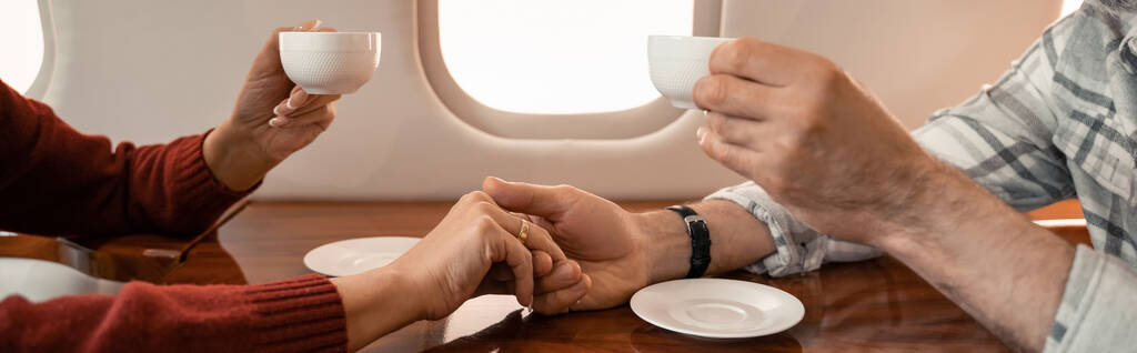 Vue recadrée du couple mature avec des tasses tenant la main dans un avion privé, bannière  - Photo, image