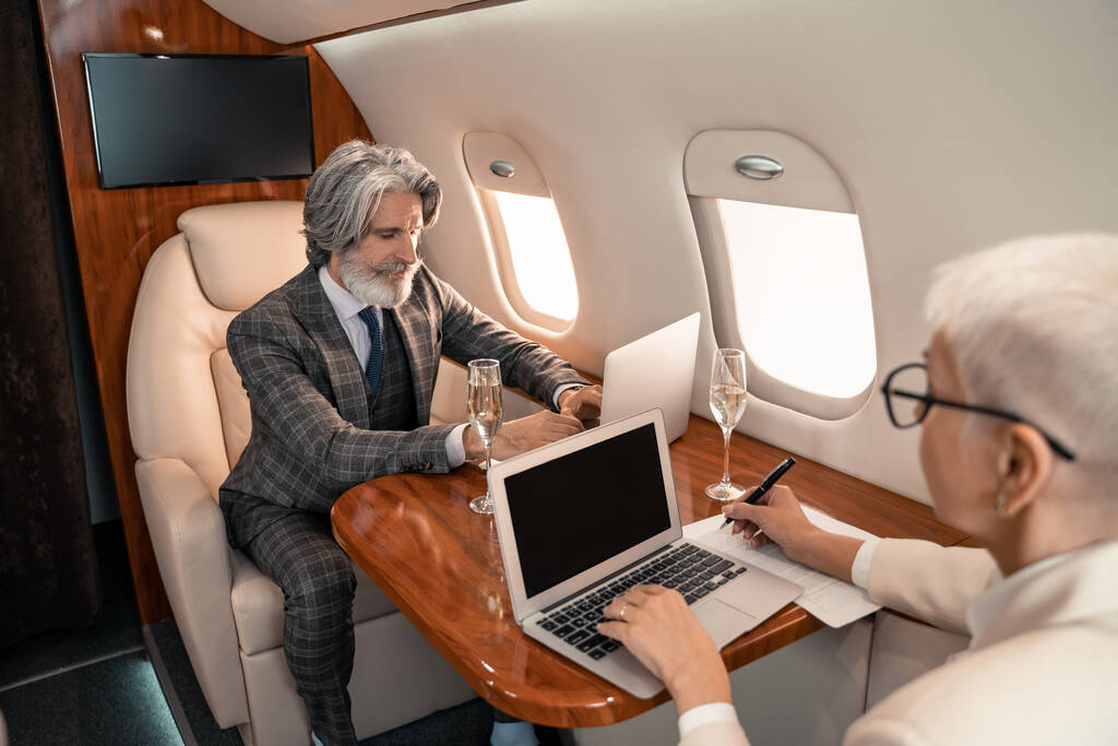 Бизнесмен, пользующийся ноутбуком рядом с размытой деловой женщиной, пишущей на бумаге и шампанским в частном самолете  - Фото, изображение
