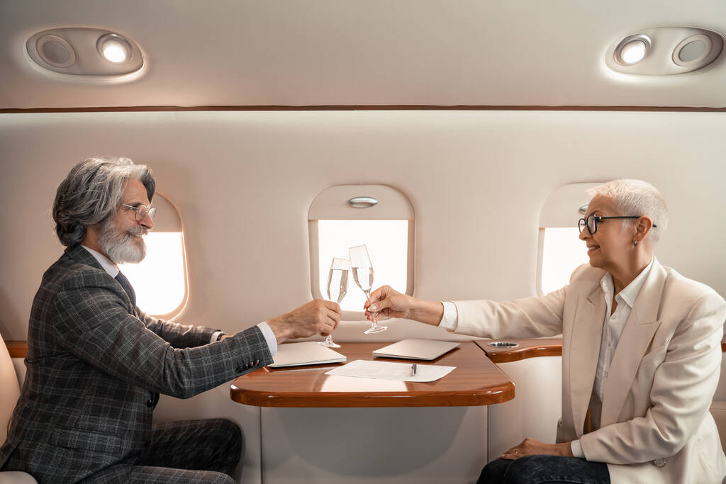 Zijaanzicht van lachende zakenmensen die met champagne aankloppen in de buurt van laptops in een privéjet  - Foto, afbeelding