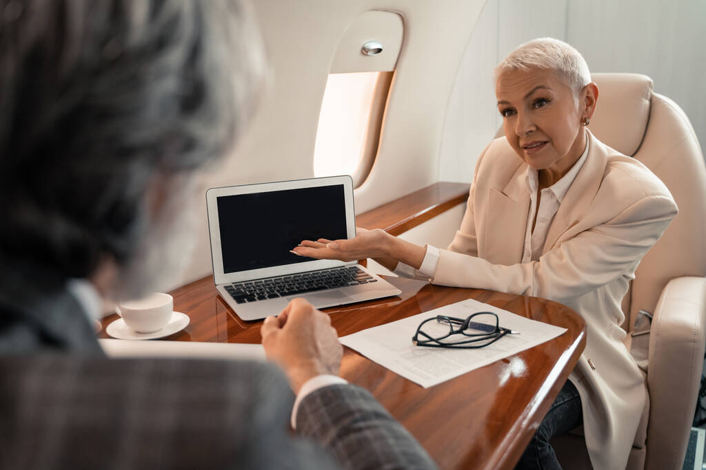 Geschäftsfrau zeigt mit der Hand auf Laptop in der Nähe von Dokument und Geschäftsmann im Flugzeug  - Foto, Bild