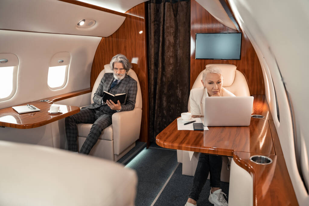 Mujer de negocios utilizando el ordenador portátil cerca de café y socio con portátil en avión privado  - Foto, Imagen
