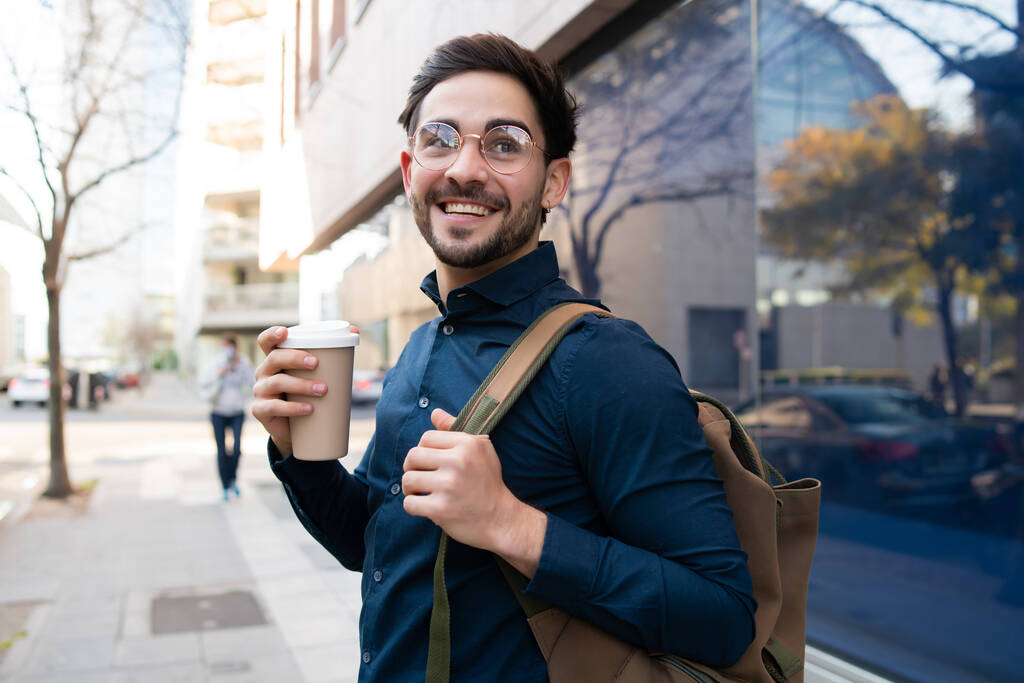Νεαρός κρατώντας ένα φλιτζάνι καφέ, ενώ περπατάτε έξω. - Φωτογραφία, εικόνα