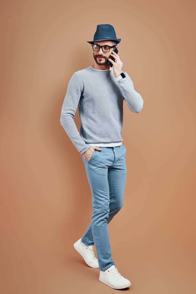 Normal giyinen genç bir adam, kahverengi arka planda dikilirken akıllı telefonla konuşuyor. - Fotoğraf, Görsel