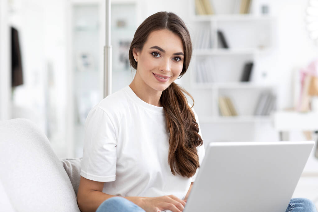 Doğal bir gülüşü olan güzel bir kız evdeki oturma odasında rahat bir koltukta dizüstü bilgisayarla oturuyor. Arkadaşlarla çevrimiçi bağlantı kur. - Fotoğraf, Görsel