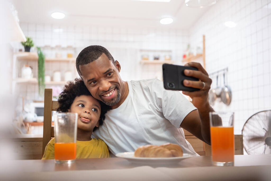 Gelukkig glimlachende Afro-Amerikaanse vader en kleine jongen nemen selfie met smartphone samen in de keuken. Vrolijke familie doet videogesprek op mobiele telefoon thuis. - Foto, afbeelding